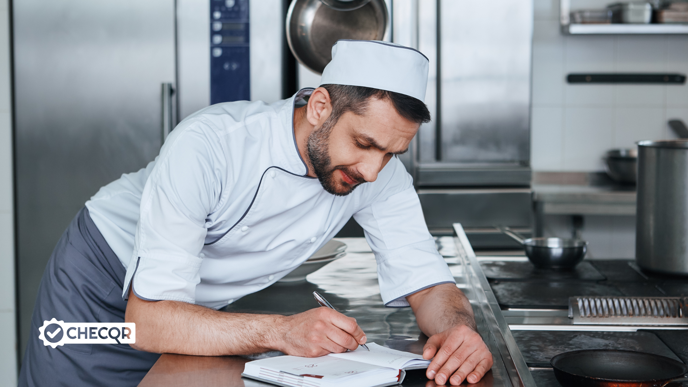 Man in chefs whites and a blue apron leaning on a commercial kitchen counter writing in a book